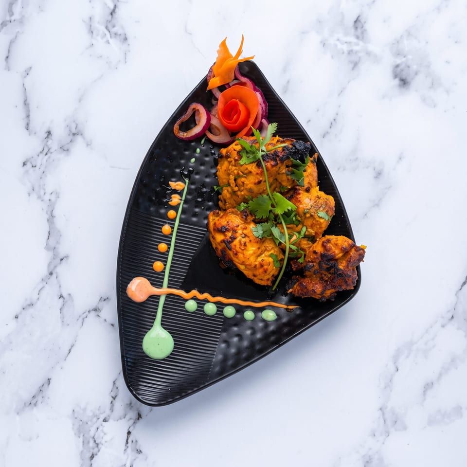Grilled chicken tikka with cilantro, onion, and carrot garnish on a black plate.