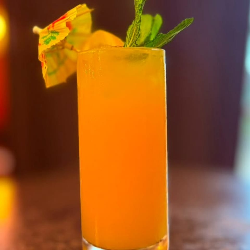 Tall glass of orange cocktail with a paper umbrella, lemon slice, and mint garnish.