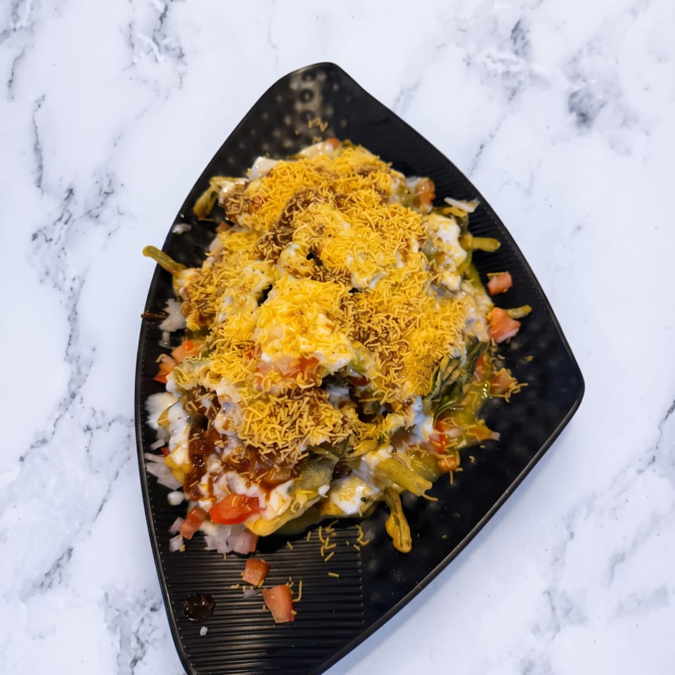 A black triangular plate of Indian chaat, topped with sev, yogurt, and chopped vegetables.