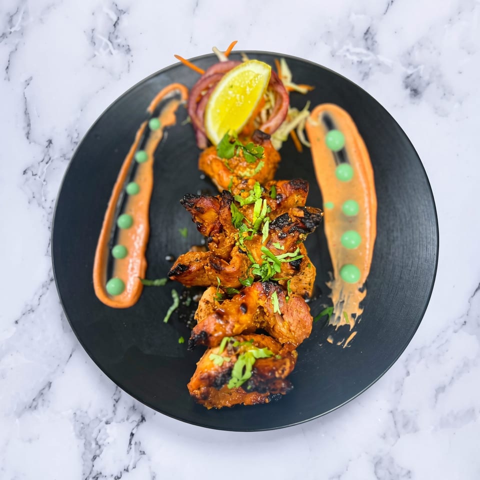 Grilled chicken tikka with cilantro, lime, red onion, and two sauces on a black plate.